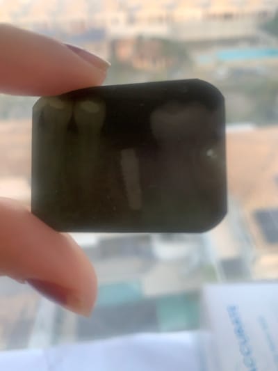 A hand, with light-colored nail polish visible on the thumb and another finger, firmly grips a dark, rectangular dental X-ray film. The film displays two distinct radiopaque dental structures resembling tooth roots or post-and-core restorations on its upper left, and a prominent, vertically oriented, threaded dental implant screw in the mid-right. The background, viewed through a window, is significantly blurred, depicting an urban landscape of multiple multi-story buildings. A light blue swimming pool is visible on a rooftop in the mid-ground, and dark photovoltaic solar panels are partially visible on another rooftop in the lower right. A white surface with partial blue text, including "ACCURATE," is visible in the immediate foreground below the film. This scene is situated within a modern, developed residential or commercial area of Lauro de Freitas, Brazil, observed from an elevated position.