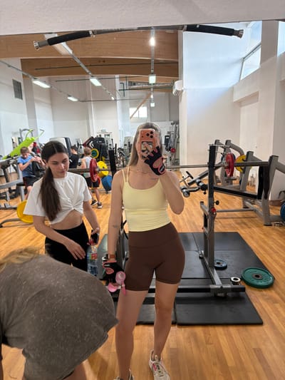 In a brightly lit gym with wooden floors, two women are visible. The woman on the right, in the foreground, is taking a mirror selfie. She is wearing a light yellow tank top and brown shorts. She has blonde hair and is wearing black and pink workout gloves. She is holding a cell phone in her right hand. To her left, another woman with dark hair is looking down and holding a water bottle. She is wearing a white knotted crop top and black shorts. In the background, several men and gym equipment, including weight racks and barbells, are visible. The gym appears to be located in Podkletnoe, Russia.