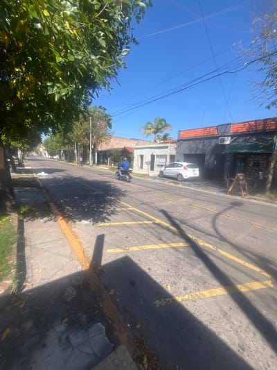 The image depicts a street in Gerli, Argentina, under a clear blue sky. A light-colored car is parked next to a dark-colored building with a red roof. Yellow stripes mark the road, which is cast in shadow. A motorcyclist is visible further down the street. Trees line the left side of the road, and a palm tree is visible in the distance.
