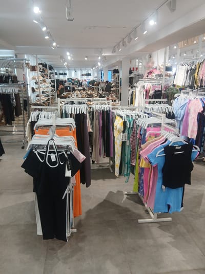 The image shows the interior of a clothing store, likely located in the city of Ávila, Spain, given the context. The store features numerous racks of clothing and shelves displaying shoes and handbags. In the foreground, several racks of clothing are visible, including a black top with white trim hanging on a hanger, a bright orange top, and other colorful garments. In the background, more clothing racks and shelves filled with various items of apparel and footwear can be seen. The store appears well-lit with multiple ceiling lights. There are no discernible people actively engaged in shopping or interaction within the frame. The environment suggests a retail setting focused on women's fashion.