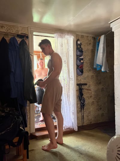 A young man, appearing to be in his late teens or early twenties, stands in the foreground facing away from the camera, looking towards an open doorway. He is shirtless and wearing light-colored shorts. In his hands, he holds a dark object and a green item, possibly a piece of clothing or a towel. Through the doorway, another young man, also shirtless, is visible, wearing dark shorts. He stands near a bottle of water on the floor. The room appears to be a rustic interior, possibly a rural house. Coats are hanging on a rack to the left. The walls are covered in patterned wallpaper. A white curtain is visible in the doorway. The floor is carpeted in a light green color. The lighting suggests it is daytime, with sunlight streaming through the doorway. There is a white structure on the right side of the frame, possibly a stove or fireplace. The scene is candid and informal. The location is the city of Sad. Uchastki, Russia.