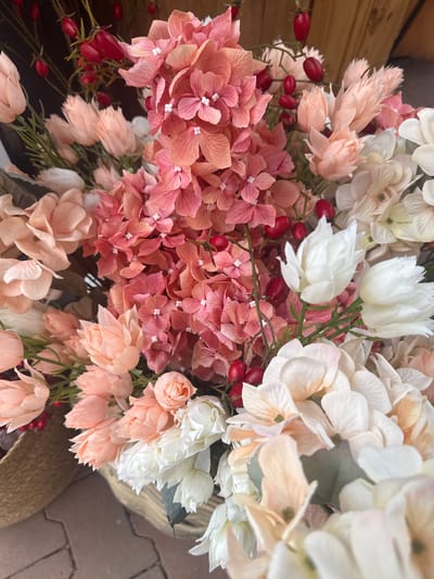This is a close-up, eye-level, vertical, indoor shot of a floral arrangement displayed in a woven basket. The arrangement features a mix of artificial flowers and red berries. Dominant floral colors include various shades of pink and peach, alongside white and cream-colored blossoms. The textures vary from the delicate, layered petals of some flowers to the more rounded clusters of others. Small, bright red berries are scattered throughout the bouquet on thin stems. The background is dark and slightly out of focus, with hints of a wooden surface and a neutral floor surface that appears to be made of interlocking pavers. There are no people present in the image, and no visible context to suggest a specific location within Fritzlar, Germany, other than what is provided in the prompt.