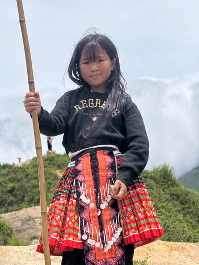 A young Asian girl, likely of ethnic minority descent from Bắc Yên, Vietnam, stands outdoors holding a bamboo pole. She wears a black hoodie with gold lettering and a vibrant, pleated skirt adorned with intricate patterns and silver embellishments. The background features a hilly, vegetated landscape under a cloudy sky. The girl is positioned in the foreground, looking towards the camera with a neutral expression.