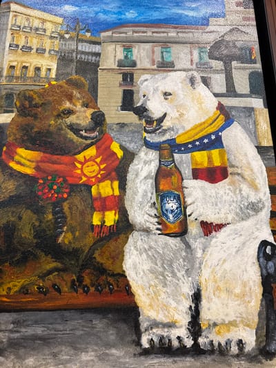 The painting depicts two anthropomorphic bears, one brown and one white, sitting on a bench outdoors. Both bears are wearing scarves. The brown bear has a red and yellow checkered scarf with a sun motif and a circular green and red decoration. The white bear wears a blue scarf with yellow stripes and white stars, and a red and yellow striped scarf underneath. The white bear is holding a bottle of "Polar" brand beer. In the background, there are buildings with balconies and windows, suggesting an urban setting. The sky is blue with clouds. The scene appears to be a stylized representation of a city square, possibly in Torrejón de Ardoz, Spain, given the context provided. The overall impression is whimsical and festive.