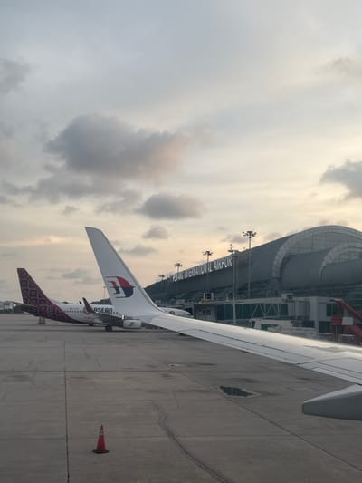 The image shows an airplane parked at Penang International Airport. The tail of the plane is visible with the Malaysia Airlines logo. The airport building is in the background. A safety cone is on the tarmac near the plane. The sky is cloudy. Penang International Airport is located in George Town, Malaysia.
