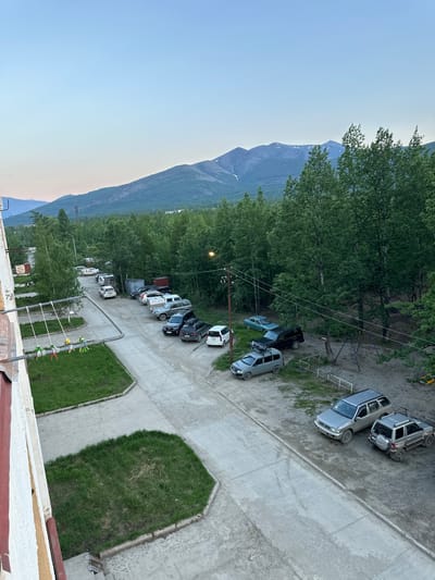 This is a high-angle, full-shot, outdoor, daytime photograph taken in the city of Sinegorye, Russia. The scene depicts a residential area with apartment buildings, a parking lot filled with various cars, and a backdrop of lush green trees and distant mountains. The sky is a pale blue, suggesting either early morning or late evening.
In the foreground, on the left side of the image, a portion of a cream-colored apartment building is visible, with visible signs of wear and tear on its facade, including peeling paint and some discoloration. Attached to the building is a metal structure, possibly a balcony railing, with several colorful, worn-out swing ropes hanging from it. Adjacent to the building is a patch of uneven, grassy lawn.
The main focus of the image is a paved road or driveway that curves through the scene. A row of parked cars lines the right side of this road. The vehicles include several SUVs and sedans, some of which appear to be older models. Notable vehicles include a silver SUV with a spare tire mounted on the back, a dark-colored SUV, and a light gray SUV. Further down the road, more cars are parked, including a white sedan.
Beyond the parking lot and road, a dense forest of tall, green trees dominates the middle ground, stretching towards the horizon. The trees are a mix of deciduous and coniferous species. In the far distance, a range of mountains with snow-capped peaks is visible against the clear blue sky. The lighting suggests it is dusk or dawn, with soft shadows and a diffused light.
The overall impression is of a somewhat rugged, natural environment bordering a developed residential area. The presence of parked cars and apartment buildings indicates a human settlement, while the extensive forest and mountains highlight the natural landscape surrounding the city. There are no visible people actively engaged in any interactions within the frame. The scene is static and quiet, with an emphasis on the built environment set against a natural backdrop.