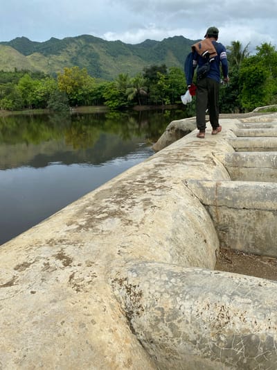 A man walks along a concrete spillway with a body of water to his left and a lush green landscape with mountains in the background. The man is wearing a dark long-sleeved shirt with white stripes on the sleeves, dark pants, a green cap, and brown shoes. He is carrying a brown jacket slung over his shoulder and a white plastic bag in his left hand. The spillway is made of rough, weathered concrete with visible cracks and stains. The water is dark and reflects the surrounding trees and sky. The landscape is characterized by dense vegetation, palm trees, and rolling green hills that rise to form mountains under a cloudy sky. The scene appears to be in a natural, possibly rural or semi-rural setting.