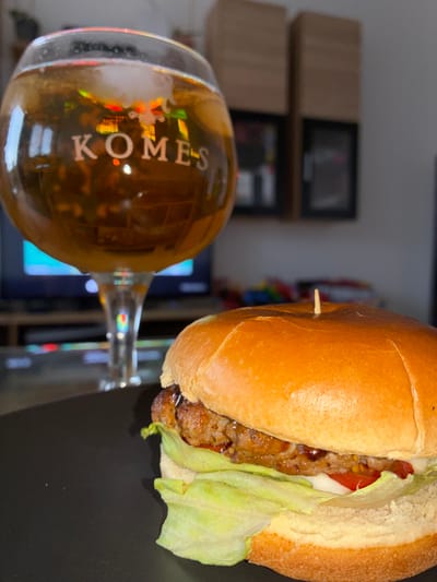 A close-up, angled shot shows a hamburger and a glass of amber liquid. The hamburger is on a dark surface, with the top bun partially covering the patty, lettuce, and a slice of tomato. A toothpick protrudes from the side of the bun. To the right, a stemmed glass filled with a golden beverage rests on its side. The glass displays text that reads "KOMES". The background is blurred, showing what appears to be a television screen with colorful reflections and some shelving units. The lighting suggests an indoor setting, possibly at night or in a dimly lit room. The overall scene conveys a casual, relaxed atmosphere, perhaps a meal enjoyed while watching television. The visible text indicates the beverage is likely a beer or similar drink. Given the location context of Toruń, Poland, "Komes" could refer to a specific brand of beer popular in the region.