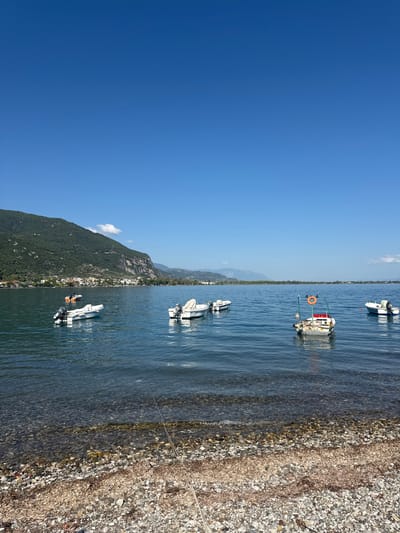 The image depicts a serene coastal scene in the Municipal Unit of Kamena Vourla, Greece. The foreground is a pebbled beach with scattered seaweed and a visible rope extending into the water. The water is clear and calm, revealing the submerged pebbles. Several small motorboats are anchored in the bay, some closer to the shore and others further out.
In the mid-ground, a line of buildings forms a coastal settlement, backed by a densely vegetated, steep mountain that dominates the left side of the frame. The mountain's slopes are covered in green foliage, with rocky outcrops visible in places. The sky is a clear, vibrant blue, with a single, small white cloud near the mountain peak.
Further in the distance, a hazy mountain range stretches across the horizon, meeting the sea. The water is a deep blue, reflecting the clear sky. The overall impression is one of tranquility and natural beauty. There are no people visible in the image.