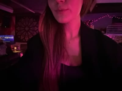 The image captures the upper torso of a female individual, from the chin downwards, in a dimly lit indoor establishment located in 마포구, South Korea. The subject's light brown hair, appearing magenta due to the ambient lighting, falls over her shoulders. She is wearing a dark-colored blazer over a dark top, with a thin gold-colored necklace visible on her neck. Her lips are partially visible. The background is characterized by strong pink and purple ambient lighting. In the upper-left quadrant, a screen displaying blue and white text or graphics is visible. Below the screen, a counter or table holds several indistinct objects, one of which emits a pink glow. Behind this area, a large, circular tapestry with an intricate, dark-patterned design is affixed to the wall. In the upper-right quadrant, a string of small, pink or purple fairy lights is horizontally strung, below which a light-colored metallic railing or frame structure is present.