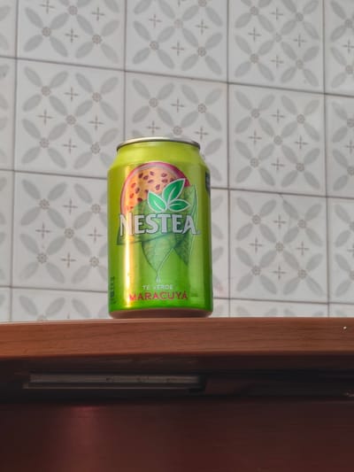 A can of Nestea green tea with passion fruit flavor stands on a brown wooden surface, likely a counter or shelf. The background is a white tiled wall with a gray, floral-like pattern repeated across the tiles. The can is green with a picture of a passion fruit and green leaves, with the words "Nestea" and "Te Verde Maracuya" prominently displayed. The image is taken indoors. There is no indication of the location being in Moaña, Spain from the image alone.