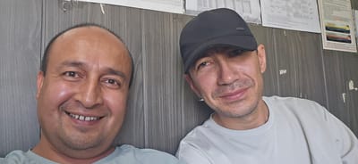 Two men are photographed from the chest up, in what appears to be an indoor setting with a grey, wood-paneled wall and some documents pinned to it in the background. The man on the left is closer to the camera, smiling broadly with his mouth open, revealing his teeth. He has dark hair and brown eyes. He is wearing a light blue t-shirt. The man on the right is wearing a black baseball cap and a light grey t-shirt. He is looking slightly to his left and has a subtle smile. He has short, dark hair and brown eyes. The lighting is moderate, with some shadows present. The overall impression is a casual selfie. The image does not provide any specific details or context that would indicate it was taken in the city of Jararyk, Uzbekistan.