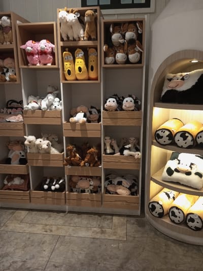 This image depicts an interior retail or display space located in Tân Kỳ Commune, Vietnam. The scene is dominated by a multi-tiered display system filled with numerous plush toys and merchandise.
On the left and center, a large white shelving unit features multiple wooden cubbies/crates, each containing various plush items. These include white sheep/goats, yellow caterpillar-like figures, pink sheep, brown horses, white bears or dogs, and numerous black and white cow-themed plush toys and pillows, some equipped with small bells.
To the right, a separate, curved display unit with light wooden shelves is present. This unit features integrated warm downlighting. It displays black and white cow-patterned items, including flat pillows/cushions on upper shelves and cylindrical, rolled blankets or pillows on lower shelves.
The floor is a light grey concrete or tiled surface with visible seams. The background wall is white, with a dark, rectangular element visible near the top center. No people are present in the scene.