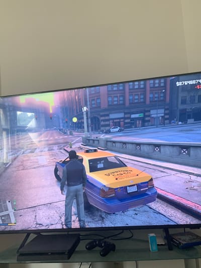The image shows a screenshot from a video game, likely Grand Theft Auto V, displayed on a large television screen. The game environment depicts a city street scene with a taxi cab in the foreground. A male character, dressed in a grey vest over a dark long-sleeved shirt and grey pants, stands behind the taxi. The taxi is yellow and purple with "DOWNTOWN CAB CO" written on its rear. A license plate reading "8SVZ130" is visible on the taxi. In the background, there are buildings, streetlights, traffic signals, and a police car. The top right corner of the screen displays in-game currency amounts, "$27646074" and "-$7". The scene appears to be set during dusk or dawn, with warm lighting casting long shadows. The television is placed on a stand below which a game console and controllers are visible, along with some boxed games. There is no indication that this scene is related to the city of Vejprty, Czechia.