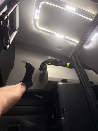 A person's leg, wearing a dark sock, is propped up on a surface inside a vehicle. The leg is positioned from the lower thigh to the foot. Above the leg, the interior of what appears to be a truck cab is visible, featuring ceiling lights and a folded garment with reflective yellow detailing draped over a storage unit. A black curtain hangs to the right. In the background, part of an extendable ladder is mounted vertically on the left side. The lighting in the scene is artificial, with bright overhead lights and some green indicator lights visible lower down. The overall environment suggests the interior of a commercial vehicle, possibly a truck, in a setting that is not specifically identifiable as New Britain, United States of America.