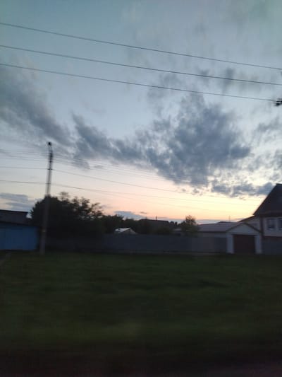 The image depicts a dusky sky with scattered clouds over a rural landscape. Three parallel power lines stretch horizontally across the upper portion of the frame. In the mid-ground, a residential area is visible, featuring houses and a fence. A lone utility pole stands to the left of center, with power lines extending from it. The foreground is dominated by a grassy field. The scene appears to be located in the village of Barabanovka, Russia, given the context provided, and suggests an outdoor setting at twilight.