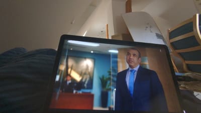 The image shows a laptop computer open on a bed in a room with a white ceiling and white walls. The laptop screen displays a scene from a television show, with a man in a blue suit and a blue tie standing in an office setting. The man has a serious expression on his face. There is a plant visible in the background, and the office appears to be modern and well-lit. The room is cluttered with furniture and blankets, and the bedspread is blue. The laptop is in focus, and the rest of the scene is blurred. This room is located in Schwabach, Germany, a town in the Bavarian region of Germany. The exact location of the image cannot be pinpointed with the given coordinates, but it is within the city of Schwabach.