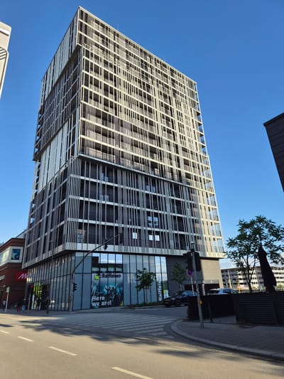 The image presents a modern, tall building under a clear blue sky, situated at a city intersection on a sunny day. The multi-story structure features a distinctive architectural design with a facade composed of numerous vertical, light-colored slats that partially enclose balconies equipped with dark metal railings. These slats create a visually striking, repetitive vertical pattern across the building's exterior.
The ground floor of the building is dedicated to commercial spaces with large glass windows. A prominent "Raiffeisen" bank branch occupies a corner, displaying its green logo and a large window advertisement featuring a group of people and the slogan "Here we are!". Adjacent to the bank, a partial red sign for "Möbel" is visible. To the immediate left of the main building, a lower, reddish-brown building with a white star logo on a blue background and a red sign starting with "M" is visible.
In the foreground, a paved street with a clearly marked pedestrian crosswalk and white dashed lines is visible. A dark grey traffic light pole with a signal box stands to the right of the crosswalk. A black SUV and a white SUV are seen parked or maneuvering near an entrance to an underground garage further back on the right side of the street. The sidewalk is paved with light-colored slabs, and several pedestrians are visible walking in the lower-left section of the image. A small tree with green leaves stands near a dark wooden fence on the right side of the sidewalk. Shadows from the buildings and street furniture are cast onto the street and sidewalk, indicating bright sunlight. The scene is located in Sanem, Luxembourg.