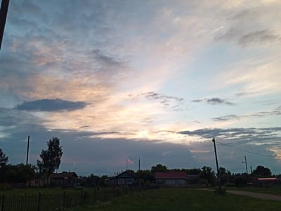 This image captures a rural landscape in Naruksovo, Russia, under a dramatic sky during what appears to be sunset or sunrise. The foreground is dominated by a dark green, grassy field with a black metal fence running horizontally across the bottom of the frame. Behind the fence, several houses are visible, varying in style and condition. On the left, a light-colored house with a dark roof is partially obscured by trees. Further right, a dark green house with white-trimmed windows stands out, followed by a smaller house with a red roof. Trees and other vegetation are scattered throughout the scene, forming a treeline behind the houses. Two utility poles with wires are prominent features in the mid-ground. The sky above is filled with a complex cloud formation, with patches of light blue, grey, and soft pink and orange hues suggesting the presence of the sun below the horizon. The overall impression is one of a quiet, pastoral setting at the transitional hours of the day.