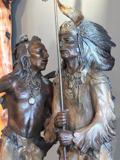 A close-up shot of a sculpture depicting two Native American figures. The top figure, adorned with a feathered headdress, holds a pole. Below, another figure lies seemingly lifeless. Both sculptures feature intricate details of clothing and facial expressions, emphasizing the weight of the pole. The sculpture's material appears to be bronze or a similar metal, giving it a rustic, textured look. The lighting emphasizes the depth of the figures.