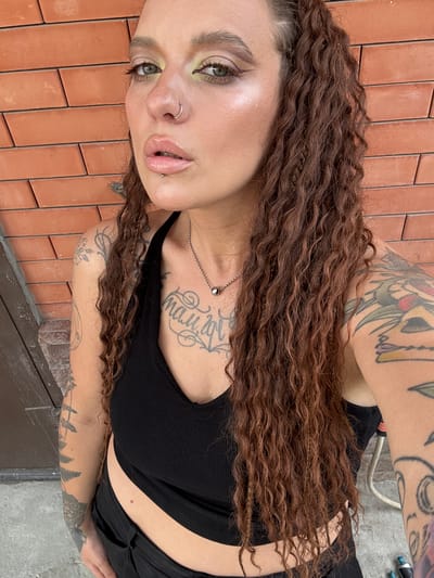 The media file is a portrait of a young woman with long, curly brown hair. She has numerous tattoos visible on her arms and chest, and she is wearing a black crop top and black pants. She has striking green eyes and is wearing makeup, including eyeshadow and lip gloss. She has piercings on her nose and lip. The background shows reddish-brown brickwork, suggesting an outdoor setting, possibly a wall or patio. The lighting appears natural and suggests it is daytime. There is no visible text in the image. The woman's expression is neutral, and the overall mood is casual and personal. The location context is Grushevskaya, Russia.