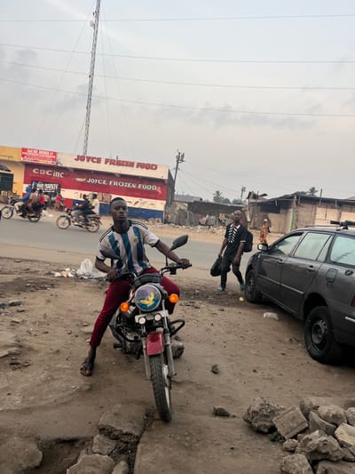 A man in an Argentina soccer jersey sits astride a red motorcycle on a dusty roadside. Next to him is a dark-colored station wagon. Behind him, two people in black are walking away from the camera. In the background, there are people on other motorcycles in front of a building called Joyce Frozen Food. An antenna tower and power lines are visible. The ground near the motorcycle is littered. Rocks are piled on the right side of the image. The sky is overcast.