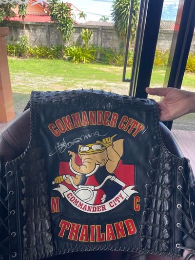 A black leather motorcycle vest, featuring genuine crocodile skin inserts on the shoulders and side panels, is displayed prominently in the foreground. The vest's central back design includes a red patch with yellow-outlined text "COMMANDER CITY" across the top. Below this, a large embroidered emblem depicts a cartoon elephant wearing yellow goggles and a red shirt, riding a white motorcycle, set against a red rectangular background. A white banner below the elephant reads "COMMANDER CITY" in black text, flanked by a red "M" on the left and a red "C" on the right. A yellow-outlined red text patch at the bottom reads "THAILAND." A white handwritten signature is visible on the upper left side of the vest. The vest features prominent leather lacing with silver-colored eyelets along the right side. An unidentified human right hand is visible on the right, lightly grasping the upper right side of the vest.

The scene is set indoors, looking out through a window. The background reveals a sunlit residential garden in Ban Yid, Thailand. A vibrant green lawn occupies the immediate background, transitioning to a grey concrete privacy wall running horizontally across the mid-ground. Beyond the wall, various tropical plants, including palm trees and broad-leafed foliage, are visible. A house with an orange or red-tiled roof is partially visible in the upper left background. The window frame appears to be dark-colored metal. The overall lighting suggests clear daylight conditions.