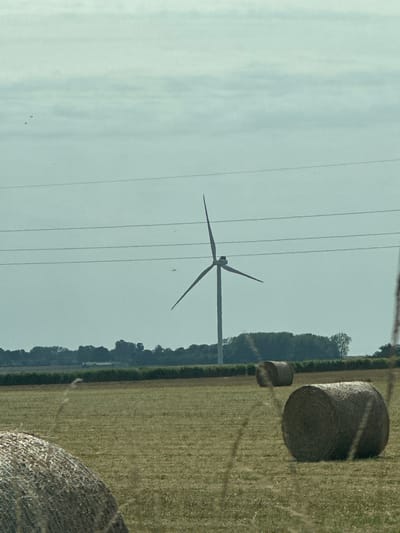 The image depicts a wide shot of a rural landscape. In the foreground, several large, round hay bales are scattered across a dry, yellowish-brown field. Some of the bales are in focus, showing their tightly wrapped texture, while others are slightly out of focus. In the middle ground, a solitary wind turbine stands tall against a pale blue, cloudy sky. Its three blades are visible, one extended upwards and the other two angled downwards. A series of thin, dark power lines stretch horizontally across the sky, intersecting in front of the wind turbine. The background features a dense line of dark green trees, forming a natural horizon. Beyond the trees, faint structures can be observed, suggesting the presence of a small settlement or farm. The overall scene is tranquil and evokes a sense of open space and agricultural activity. The location is identified as Longueil, France.