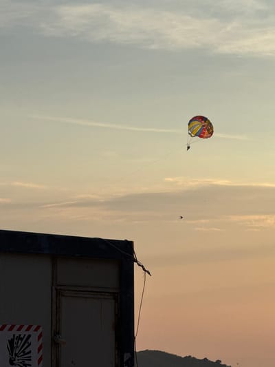 In Vrijesno, Montenegro, at sunset, a parasailer floats in the sky above a dark structure and a distant hillside. The parasail is multi-colored, with sections of red, yellow, green, and blue. Below the parasail, two small figures can be seen, presumably the parasailer and the tow boat operator. A thin line connects the parasail to an unseen tow boat. The sky is a gradient of soft orange and pale blue, with wispy clouds and faint contrails. In the foreground, the top of a dark, utilitarian structure is visible, its roofline dark against the sky. The structure appears to be made of metal and has a weathered door. A caution sign with red and white stripes and an explosion symbol is partially visible on the left side of the structure. The hillside in the distance is silhouetted against the setting sun, with a few trees visible along its crest. The overall scene is tranquil, with the vibrant parasail providing a focal point against the muted colors of the sky and landscape.