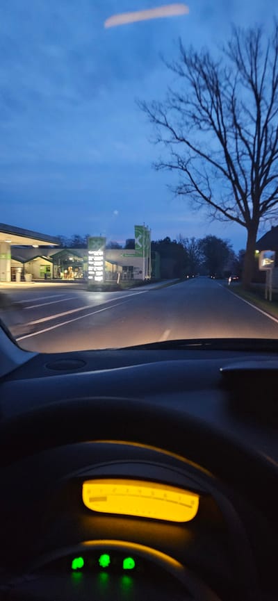 The scene depicts a forward-facing view from inside a vehicle at dusk or night, positioned on a road approaching the Aral gas station at Gartröger Weg 1, 33397 Rietberg, Germany. The sky is dark blue with minimal cloud cover.

In the foreground, the dark-colored dashboard of the vehicle is visible, along with the top portion of a steering wheel. The instrument cluster displays a horizontal, yellow-illuminated segment with numerical markings, and three distinct, circular green indicator lights are illuminated below it. The vehicle's headlights cast a warm light onto the asphalt road ahead, revealing white lane markings.

On the left side of the road, the Aral gas station complex is brightly lit. It includes a multi-columned canopy structure, a building housing a convenience store with illuminated interiors and exterior signage. A tall, vertical price display sign is prominently illuminated. Three green flags, each bearing the white Aral 'X' logo, are visible outside the building, which also has "REFINER" text on its facade.

On the right side of the road, a large, bare-branched deciduous tree stands prominently against the dark sky. Further down the road, a reflective roadside marker is partially visible. The road extends into the distance, where a single red taillight from another vehicle is faintly visible. No individuals are discernibly present in the image.