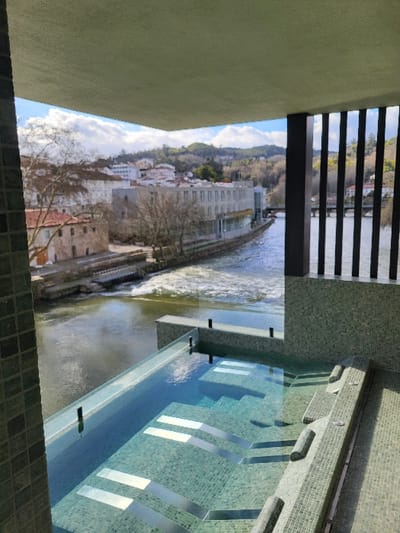 This image presents a panoramic outdoor view from a private balcony or terrace in São Pedro do Sul, Portugal, overlooking a river and the town.
In the foreground, a rectangular infinity-edge pool or jacuzzi is positioned, approximately 2-3 meters in width. The pool's interior is finished with light green mosaic tiles, featuring multiple submerged stainless steel steps and benches. A clear glass barrier forms the edge of the pool, providing an unobstructed view of the scene beyond. To the left, a wall covered in light green mosaic tiles is partially visible. To the right, a dark, vertically slatted balustrade acts as a partial enclosure. A flat, light-colored concrete slab forms the ceiling of the overhead structure.
Beyond the pool, a river flows from left to right across the midground. A prominent section of turbulent, white water, indicative of a weir or rapids, is visible in the mid-left. Along the opposing riverbank, various buildings are present. On the left, an older stone building with a red-tiled roof stands near the water's edge. Further right, a larger, modern multi-story building with extensive glass facades is situated directly on the riverbank. In the distance, a multi-arched bridge spans the river.
The background comprises gently sloping hills, predominantly covered with bare deciduous trees, suggesting a winter or early spring season. Scattered white buildings belonging to São Pedro do Sul are visible throughout the landscape. The sky is partly cloudy with visible patches of blue. No individuals are present in the scene, and no specific actions or interactions are depicted.