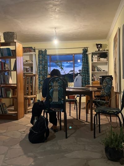 The image depicts an indoor scene in Chitungwiza, Zimbabwe, illuminated by artificial overhead light during what appears to be twilight or night. One individual, seen from behind, is seated on a patterned chair (blue, yellow, black geometric with a lighthouse graphic) at a dark wooden rectangular table. The person wears a dark long-sleeved top and dark trousers; their dark hair extends to the shoulders. A black backpack with a silver water bottle is positioned on the flagstone floor beneath their chair.

The room features a large window at the rear, revealing a dark blue sky with clouds and a prominent, spherical bright light source, consistent with a moon. The window is flanked by curtains with a blue, yellow, and black geometric pattern. To the left, a tall, light-colored wooden bookshelf filled with books stands, topped with a woven basket. To the right, a white multi-shelf unit holds books, boxes, and a decorative wooden clock. A large vertical artwork hangs on the right wall.

On the table, a large translucent white plastic jug, papers, and an electronic device or printer are visible. Cables extend from the table area. The floor is made of irregularly shaped flagstone tiles. A single bare lightbulb hangs from the textured, light-colored ceiling. Cream-colored walls with white crown molding complete the interior. A potted succulent plant rests on the floor in the lower right corner.