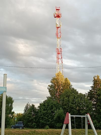 A tall, red and white telecommunications tower dominates the center of the frame, reaching towards a cloudy sky. The tower is segmented and has multiple platforms with antennae. In the foreground, on the right side of the image, a children's swing set with red and white supports is visible. To the left, a blue and white directional sign is partially visible, with some Cyrillic text that reads "ЕНИЕ". Below the sign and to the left of the tower, a dark-colored SUV with a roof rack is parked on a grassy area, with a yellow pedestrian crossing sign visible behind it. Dense green trees form a backdrop behind the swing set and the SUV, with some trees near the tower catching the golden light of what appears to be late afternoon or early evening. The overall scene is one of a semi-rural or suburban setting, with a mix of infrastructure and recreational elements under a dramatic, overcast sky. The time of day is likely late afternoon or early evening, indicated by the lighting on the trees, and the weather appears to be cloudy. The location is Smolino, Russia.