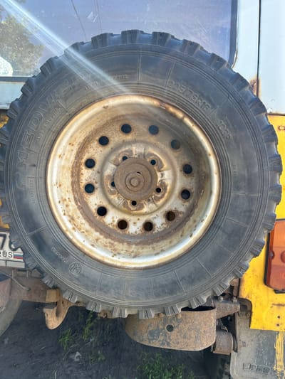 A close-up, eye-level, vertical shot shows a spare tire mounted on the rear of a weathered, yellow vehicle, likely a utility vehicle or truck, which is situated in an outdoor environment. The spare tire itself is black with a silver-colored rim that exhibits significant rust and pitting. Visible tire markings include "MEDVED STEEL RADIAL" on the left side and "225/75R16 103P" on the right side. The rim has multiple bolt holes and a central hub assembly. The vehicle's bodywork appears faded and possibly repainted, with patches of rust visible, particularly around the mounting bracket for the spare tire. A small portion of a license plate with Russian text and a flag is visible at the bottom left. The background includes the vehicle's glass window, reflecting sunlight and some greenery. The ground beneath appears to be dark soil with some sparse green grass. The location is identified as Dumino, Russia. No people or animals are present in the scene.