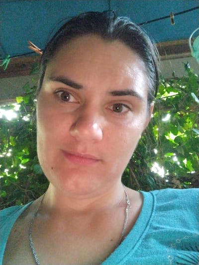This is a close-up, head-and-shoulders selfie of a woman outdoors. She has dark hair pulled back and is wearing a turquoise V-neck t-shirt with a silver chain necklace. Behind her, there are lush green leaves and branches, suggesting a natural, possibly garden setting. The lighting indicates it is daytime, with sunlight filtering through the foliage. A clothespin is visible in her hair on the left side of the frame. The woman's expression is neutral to slightly smiling, and her gaze is directed towards the camera. The overall impression is of a casual, informal moment captured in a bright, outdoor environment. There is no visible text in the image.
