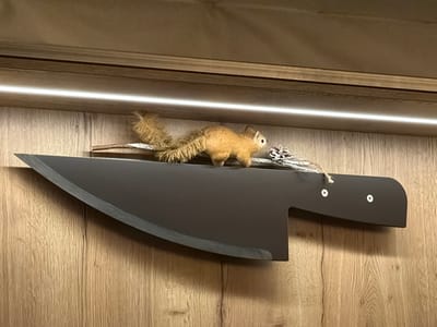A large, stylized black cleaver is mounted on a wood-grain wall beneath a recessed light. Atop the cleaver sits a small, fuzzy brown squirrel figurine with a bushy tail, alongside a pinecone and a twig. The cleaver is designed with a broad, curved blade and a rectangular handle, secured with two visible bolts. The presentation creates a whimsical contrast between the cleaver's menacing form and the squirrel's playful presence.