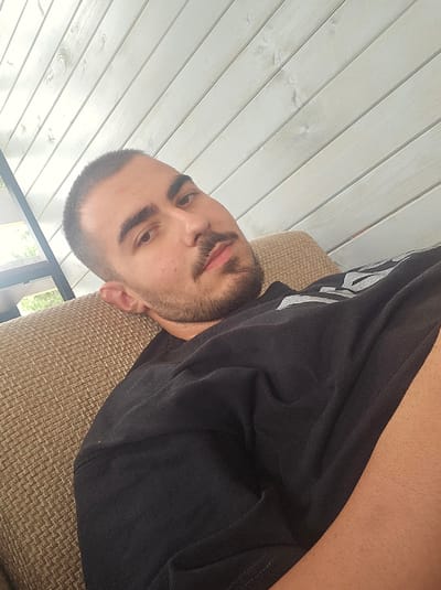 A close-up, over-the-shoulder selfie is taken from a low angle of a young man with short dark hair and a beard, resting on a textured couch. He is wearing a black t-shirt with a grey graphic design on the sleeve. The background consists of a light-colored, slatted wooden ceiling, and a partial view of outdoor greenery through a window on the left. The image appears to have been taken during daylight hours, possibly on a patio or porch.