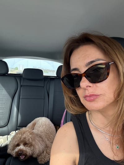 An upward-angled view captures a woman and a small dog inside a car, likely a selfie taken by the woman. The woman, positioned on the left, has shoulder-length, light brown hair. She wears large, tortoise-shell framed sunglasses that obscure her eyes, and her lips are softly parted with light pink lipstick. She is dressed in a black ribbed top and accessorized with two delicate silver necklaces, one featuring a small heart-shaped green pendant, and a ring on her finger.
To her right, a fluffy, light brown dog, resembling a poodle, rests on the black leather car seat. Its mouth is slightly open, suggesting it might be sleeping or yawning. A striped piece of fabric is visible beneath the dog.
The car's interior features black leather seats and headrests. Through the car window in the background, a bright daylight scene is visible, hinting at clear weather. The distant landscape appears open, possibly rural or suburban, but specific details are indistinct. The overall scene suggests a relaxed moment, possibly during travel in Pombal, Portugal. No visible text is present in the image.