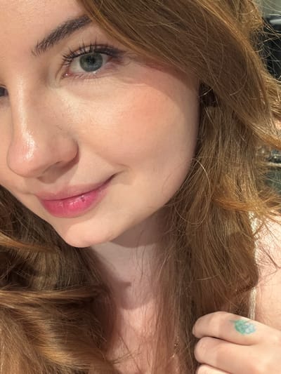 This media file is a close-up, sideways portrait of a person with long, wavy, reddish-brown hair. The person's face is partially visible, focusing on their left eye and mouth. They have a light complexion, with subtle pink blush on their cheeks and glossy pink lipstick. Their eye is light blue-green, with dark eyelashes and visible eyeliner. The eyebrow is dark and well-defined.
A finger with a small, blue and green floral tattoo is visible near the top right of the image, resting on the person's hair. The background is out of focus and appears to be an indoor setting with soft lighting. The overall mood is relaxed and intimate. There is no visible text in the image.