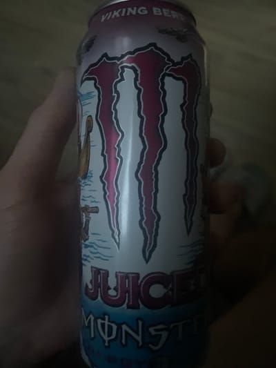 A hand is holding a can of Monster Energy drink, specifically the "Juice Monster Viking Berries" flavor. The can is primarily white with pink and dark pink accents. The prominent Monster logo is visible, along with text that reads "JUICE" and "MONSTER". The background is dark and out of focus, making it impossible to discern any specific details of the environment, including the city of Paczkowo, Poland. No people other than the implied hand holding the can are visible.