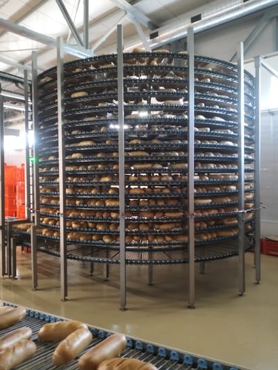 The image shows a large industrial bread cooling rack filled with loaves of bread. The rack is a spiral structure with multiple tiers, each carrying rows of bread. The bread appears to be light brown and uniformly shaped. In the foreground, a conveyor belt carries more loaves of bread. The environment appears to be a bakery or food production facility, with a concrete floor and metal structural elements visible. The lighting suggests an indoor setting. There are no people visible in the image. The image was taken in Șoldănești, Moldova.