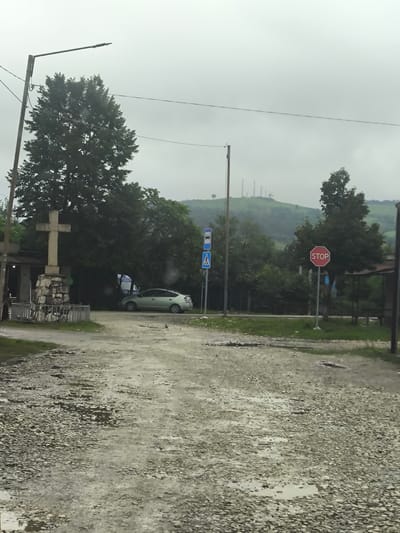 In the city of Skurdi, Georgia, on a cloudy day, a dirt road with scattered puddles leads through a rural landscape. A pale green car is parked on the right side of the road. A large stone cross, a common religious symbol, stands prominently in the foreground. Behind the cross, a dense cluster of trees creates a backdrop. Further in the distance, hills are visible, dotted with what appear to be communication towers. A stop sign and a pedestrian crossing sign are positioned near the road. The overall scene suggests a quiet, possibly remote, location.