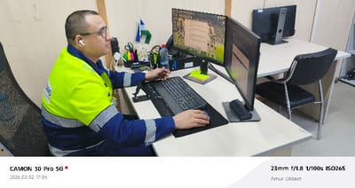 The image depicts an indoor office setting in Yukhta, Russia. An adult male is seated in a black mesh office chair at a light-colored wooden desk. He wears glasses, a neon yellow-green high-visibility vest with reflective strips over a blue long-sleeved shirt, and white wireless earbuds. His hands are positioned on a black keyboard and mouse, indicating active computer use, and he is looking towards the monitors.

The workstation includes two black computer monitors: a primary horizontal monitor displaying a desktop with a football/soccer theme and a messaging application, and a secondary vertical monitor to its right. Also on the desk are a clear plastic water bottle, a black CPU tower, a small flag resembling that of Bashkortostan, an organizer containing pens and scissors, a mobile device stand, a stack of yellow sticky notes, and a black phone handset. To the right of this workstation, another light-colored wooden desk with a black office chair and a single black computer monitor is visible. The background consists of light-colored walls, possibly partitioned, defining the workspace.