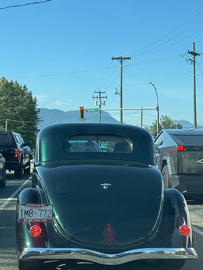 The image depicts the rear of a dark green, vintage car stopped at a red light at the intersection of Lougheed Highway and a cross street in Pitt Meadows, Canada. The car has chrome detailing and a license plate reading "1M8-772" with the "British Columbia Collector" designation. Behind the vintage car is a modern vehicle. A street sign reads "Lougheed Hwy" on the left. Power lines are visible against a clear blue sky with a mountain range in the background. The setting appears to be a sunny day. Other vehicles are also stopped behind the car.