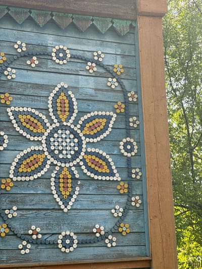 A close-up shot depicts a blue wooden fence adorned with intricate flower designs made from painted wooden spools or bottle caps. The main design is a large, stylized flower with six petals, filled with concentric circles of yellow, orange, blue, and white spools. Smaller flower motifs are scattered around, connected by lines of blue spools. The fence is topped with a wide, horizontal wooden beam, and part of a tree canopy is visible in the background. On the left side, there is a section of the fence with green and blue geometric designs. The setting suggests an outdoor location, possibly a park or yard, given the presence of the tree. The decorations on the fence give it a folk art or handcrafted appearance. The image originates from the city of Balezino, Russia.