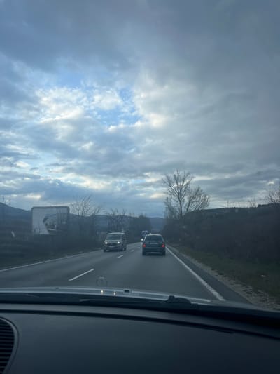 The image, captured from the interior of a vehicle displaying a Mercedes-Benz hood emblem, presents a roadway scene in or near Cherniche, Bulgaria, under an overcast sky with fragmented patches of brighter blue and white clouds. A two-lane asphalt road, appearing damp or reflective, extends into the distance, delineated by a broken white lane marker and a solid white line on the right shoulder.

Ahead, a dark-colored SUV or hatchback, possibly a Skoda, occupies the right lane, displaying illuminated taillights. Its rear license plate, partially visible, begins with "CB," indicative of Bulgarian registration. Further forward in the left lane, a silver-grey van, likely a Ford Transit model, travels with its headlights active. A third, dark-colored vehicle is partially obscured beyond the SUV.

The immediate roadside on the right features a narrow verge with sparse, bare deciduous trees and dark bushes, suggesting a non-foliage season. On the left side of the road, a large, rectangular billboard stands, displaying a damaged white surface with dark, indistinct graphics. In the background, distant hills or mountains rise on both sides, with some scattered structures visible on the right-hand slopes. No individuals are explicitly visible.
