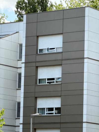 This media file shows a close-up, tilted view of a modern building's exterior. The building features a facade of alternating white and gray panels, with horizontal lines creating a segmented appearance. Several windows are visible, some with white roll-down shutters. In one of the windows, a person's head and shoulder are visible, leaning out. Trees with green foliage are seen on the left side of the frame, suggesting an outdoor setting. The sky, partially visible, is blue with some clouds, indicating daytime. A street lamp is mounted on the building's facade on the right. The lower part of the building displays more windows, some with dark interiors. The overall impression is of a contemporary residential or office building. The location is identified as Évry-Courcouronnes, France. No text is visible in the image.