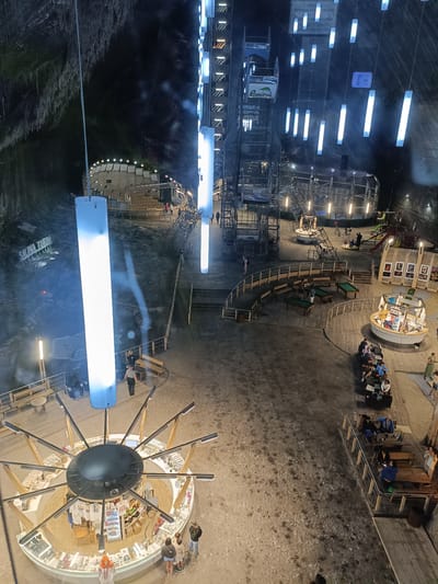 The image depicts the interior of the Salina Turda salt mine in Turda, Romania, viewed from an elevated perspective. The vast underground space is illuminated by numerous bright white LED lights, which hang from the ceiling and are integrated into large, cylindrical structures. The walls of the mine are rough and dark, showing the natural texture of the salt rock.
In the foreground, a circular kiosk with radiating arms, likely a souvenir shop, is visible. It is stocked with various items, and several people are browsing. Nearby, a tall, illuminated cylindrical structure stands prominently. Further into the mine, a recreational area is set up with pool tables, benches, and seating areas where people are gathered. A small playground with a slide can be seen in the background.
A large mining elevator or transport system is partially visible on the left side of the image, with the word "România" displayed on a banner. The ground surface appears to be a mix of sand, gravel, and possibly salt dust. Several individuals are walking around the various levels of the mine, interacting with the amenities and exploring the environment. The overall impression is of a well-developed tourist attraction within a unique natural setting.
