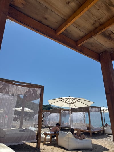This image presents a sunlit beach in Vrba, Montenegro, viewed from beneath a rustic wooden overhead structure. The top and right edges of the frame are defined by visible wooden beams and a vertical support post. The sky is a clear, bright blue, with a discernible aircraft contrail visible in the upper right quadrant.
The beach floor is composed of light-colored pebbles and sand. A series of wooden canopy beds, each featuring translucent white sheer curtains for privacy and shade, are arranged along the shoreline. Interspersed among these beds are several large, open white patio umbrellas. A blue zip tie is visible securing netting to the top left corner of the leftmost canopy structure.
Several individuals are present on the beach. In the lower right foreground, a person is seated on a white beanbag chair, looking down at an object in their lap. To their left, a shirtless male is seated at a small wooden table, interacting with items on its surface; a white towel is draped over the table. Further back, additional individuals are observed relaxing in the canopy beds, standing near the water's edge, or partially submerged in the calm, clear blue-turquoise sea. The background features green, tree-covered hills and distant white buildings lining the horizon beyond the water. The scene is illuminated by direct daylight, producing distinct shadows.
