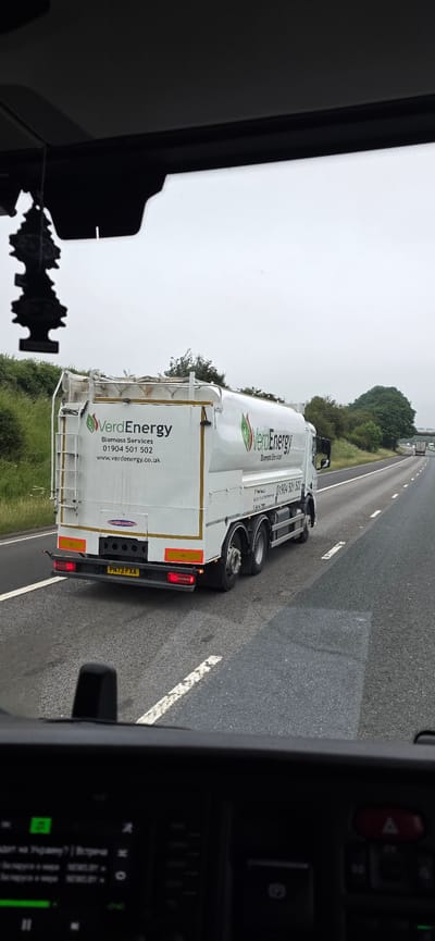 In Barton, United Kingdom, a white tanker truck with "VerdEnergy Biomass Services" branding is traveling on a highway. The truck has the license plate PN73 FXA. A black car air freshener hangs from the rearview mirror of the vehicle from which the photo is taken. The sky is overcast.